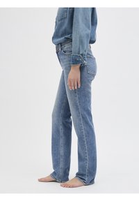 Osoba stojící naboso, oblečená v lehkých modrých denimových džínách a odpovídající denimové košili, zobrazená od pasu dolů na pozadí bílé barvy.