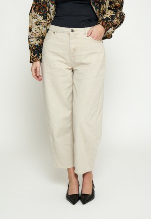 Kvinde iført højtaljede, lysebeige viddebenede jeans med sorte spidse hæle og en mønstret langærmet top, stående mod en hvid baggrund.