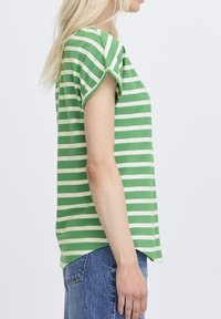 Hosszú szőke hajú nő zöld-fehér csíkos rövid ujjú inget és kék farmert visel, oldalról látható, egyszínű háttér előtt.