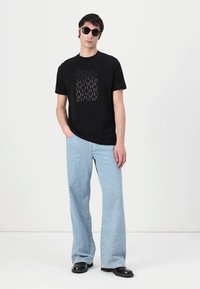 Svart bomulls T-skjorte med en teksturert grafisk design på fronten, kombinert med lyseblå vide jeans og runde svarte solbriller.