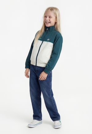 Ung pige smilende, iført en teal- og hvid lynlåstrøje, blå jeans og hvide sneakers, stående mod en ensfarvet hvid baggrund.