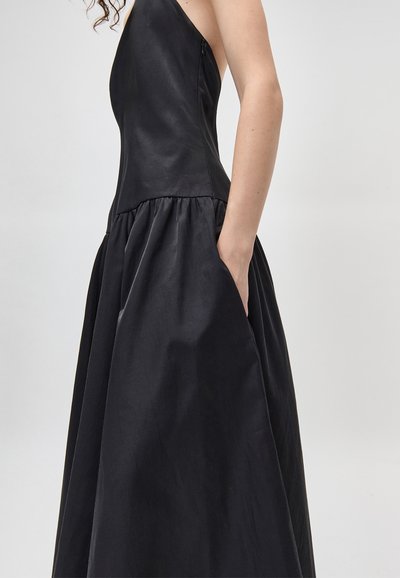 Γυναίκα που φοράει αμάνικο μαύρο φόρεμα με πλαϊνή τσέπη, τα χέρια στην τσέπη, στέκεται μπροστά σε απλό ανοιχτό γκρι φόντο.