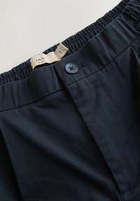 Marineblaue Slim-Fit-Hosen mit elastischem Bund, einem Knopfverschluss und sichtbarem Stoffetikett. Glatte Textur und taillierte Form.