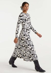 Mujer caminando con un vestido midi de manga larga en blanco y negro con estampado floral, acompañado de un accesorio para el cabello a juego y botas negras hasta la rodilla.