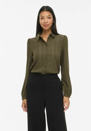 Une femme porte un chemisier vert olive boutonné avec des manches bouffantes et un pantalon noir à jambes larges sur un fond blanc uni.
