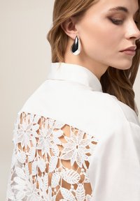 Weißes Hemd mit kunstvollen floralen Spitzenverzierungen auf dem Rücken, kombiniert mit glänzenden schwarzen Tropfenohrringen, die eine klare Ästhetik hervorheben.