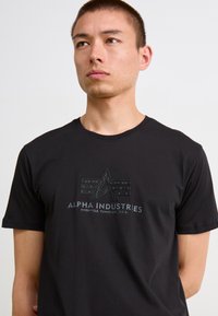 Svart bomull T-skjorte med en teksturert logo design i midten. Logoen inkluderer teksten "ALPHA INDUSTRIES" og geometriske aksenter.