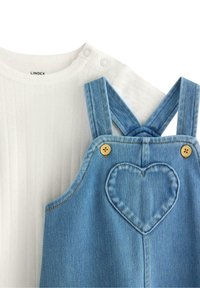 Weißes, geripptes kurzärmliges Hemd mit Druckknöpfen neben blauen Jeanslatzhosen mit herzförmiger Stickerei und Holzknöpfen.