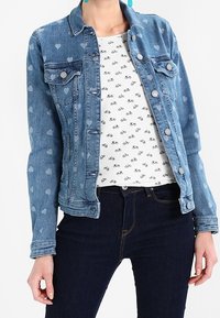 Jeansjacka med hjärtmönster, ljusblå färg, med två bröstfickor och silverknappar, lagrad över en vit t-shirt med cykeltryck.