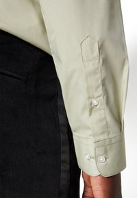 Chemise à manches longues vert clair avec un poignet pointu et deux boutons en perle, associée à un tissu noir foncé. Finition textile lisse.