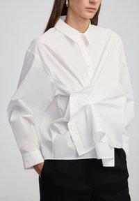 White button-up shirt made of smooth fabric featuring a draped design at the waist, long sleeves, and a classic collar. Worn with black pants.