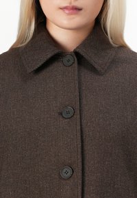 Femme portant un manteau marron texturé boutonné avec un large col, ne montrant que le bas du visage et le haut du torse.