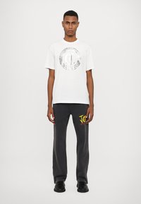Hvid bomulds t-shirt med et rundt sølv grafisk design, parret med løse sorte sweatpants med gul tekst og en afslappet pasform.