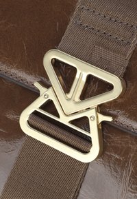 Borsa in pelle marrone con una finitura lucida. Presenta una fibbia in metallo dorato con design geometrico e una tracolla intrecciata marrone attaccata.