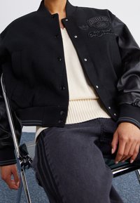 Zwart varsityjack met een wollen lijf en leren mouwen. Heeft geborduurde logo's, drukknoopsluitingen en geribde manchetten. Draagt over een witte sweater.