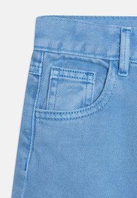 Světle modrá džínovina s pasem, malou přední kapsou a prošívanými detaily. Textura je hladká s klasickým jeanovým povrchem.