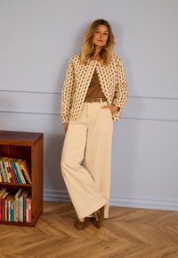 Žena stoji s rukom u džepu, nosi cvjetnu jaknu, smeđu majicu, široke bež hlače i smeđe cipele pored drvene police za knjige.