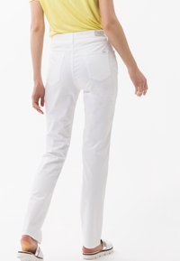 Vita raka byxor med bakfickor och en klassisk midja, kombinerade med en gul topp och silverfärgade sandaler, framför en vit bakgrund.