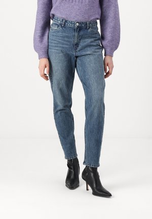 Henkilö pukeutuneena korkeavyötäröisiin sinisiin farkkuihin, mustiin teräväkärkisiin nilkkureihin, joissa on korko, ja violettiin pitkähihaiseen neulepuseroon, seisomassa valkoisella lattialla.