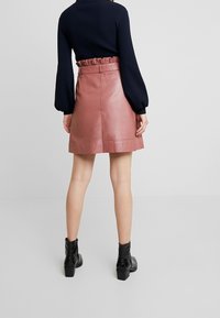 Naisen vaaleanpunainen nahkairtopaita, jossa on rypytetty vyötärö, takasaumadetaillia, yhdistettynä tummaan pitkähiharaiseen puseroon ja mustiin nilkkabootseihin.