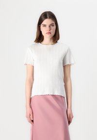 Hvit kortermet topp med teksturerte vertikale striper, bølget kant og rund hals, båret over en rosa skjørt.