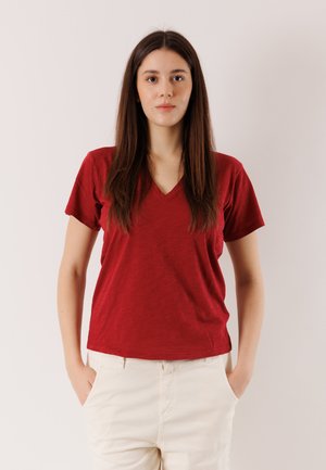 Donna con capelli lunghi castani, che indossa una T-shirt rossa con scollo a V e pantaloni bianchi, in piedi con le mani in tasca contro uno sfondo chiaro uniforme.