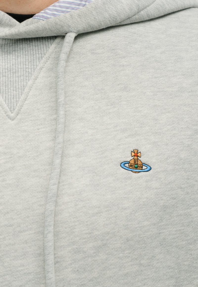 Sweat à capuche gris avec un col côtelé texturé. Présente un petit motif brodé d'un bateau avec un anneau bleu et un détail en forme de croix.