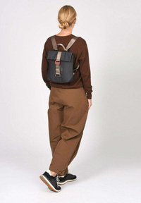 Mochila negra con solapa plegable, correas grises y herrajes rojos. Se lleva en la espalda, combinada con pantalones marrones y zapatillas negras.