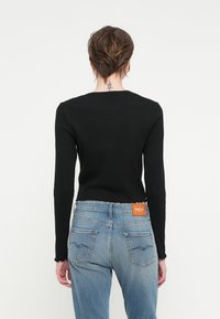 Črna rebrasta majica z dolgimi rokavi v kombinaciji s svetlo modrimi kavbojkami z raztrganim robom in zadnjimi žepi. Oznaka vidna na pasu kavbojk.