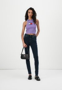 Purple ribbed tank top with a logo patch, dark denim skinny jeans, black handbag, and black loafers, showcasing form-fitting design and textures.