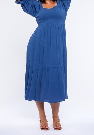Blue midi dress with a smocked bodice, puff sleeves, and a tiered skirt. Made of lightweight fabric with a smooth texture. Brown sandals worn.