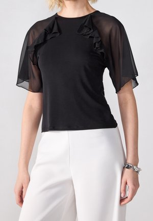 Czarny top z krótkimi, przezroczystymi, falbaniastymi rękawami i dopasowanym krojem. Materiał jest gładki z lekkim połyskiem, zestawiony z białymi spodniami.