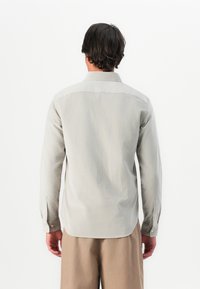 Man with dark hair wearing a light grey long-sleeve shirt and beige pants, facing away against a plain white background.