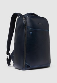 Marineblauwe rugzak gemaakt van glad materiaal met een strakke uitstraling, voorzien van een voorzak met rits, gewatteerde schouderbanden en een subtiele logo.