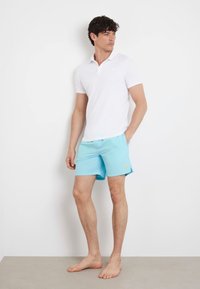 Polo shirt bianco a maniche corte da uomo, abbinato a pantaloni da bagno azzurri chiari con un piccolo logo, su uno sfondo neutro.