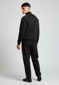 Tuta nera con zip, collo alto e polsini a coste, dal design elegante, tessuto liscio, e pantaloni sportivi coordinati con vestibilità atletica.