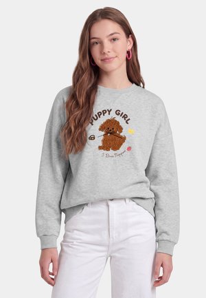 Giovane donna con lunghi capelli castani indossa una felpa grigia con un cucciolo marrone e il testo "Puppy Girl I Love Puppies,", abbinata a pantaloni bianchi.