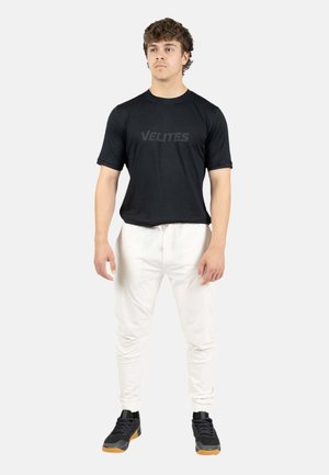 Joven de pie con una camiseta negra "VELITES", pantalones jogger blancos y zapatillas negras con suelas de goma sobre un fondo claro y sencillo.