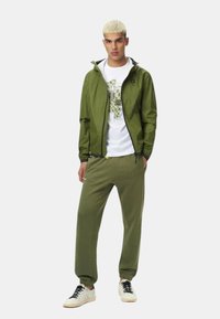 Giacca verde oliva con cappuccio, t-shirt grafica bianca, pantaloni jogger oliva e sneakers bianche. Materiale liscio, design semplice, vestibilità rilassata.