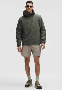 Groene capuchonjack met een gestructureerd oppervlak, een rits aan de voorkant en twee borstzakken, gecombineerd met lichtbeige shorts en bruine sneakers.