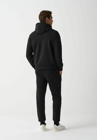 Sudadera negra con un corte holgado y capucha ajustable, combinada con pantalones de chándal a juego. Completa con zapatillas blancas. Textura suave.