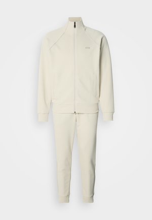 Bela športna jakna z zadrgo, v kompletu s hlačami, ki ima gladko teksturo, rebraste manšete, bočne žepe in minimalističen logotip.