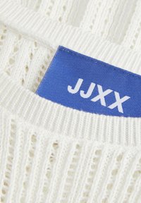 Hvit strikket genser med ribbestruktur og åpne strikkemønstre. Blå etikett med "JJXX" i hvitt, plassert nær kragen.