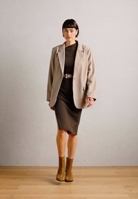 Beige blazer over a dark ribbed dress, styled with a light belt. Brown ankle boots. Smooth texture, tailored fit, and classic design.