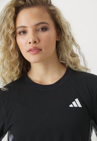Svart atletisk skjorte med korte ermer, rund halslinje, med hvit Adidas-logo på venstre bryst og teksturert stoff.
