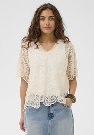 Young woman with wavy brown hair wearing a cream lace top and light blue denim skirt, with a green bangle on her wrist.