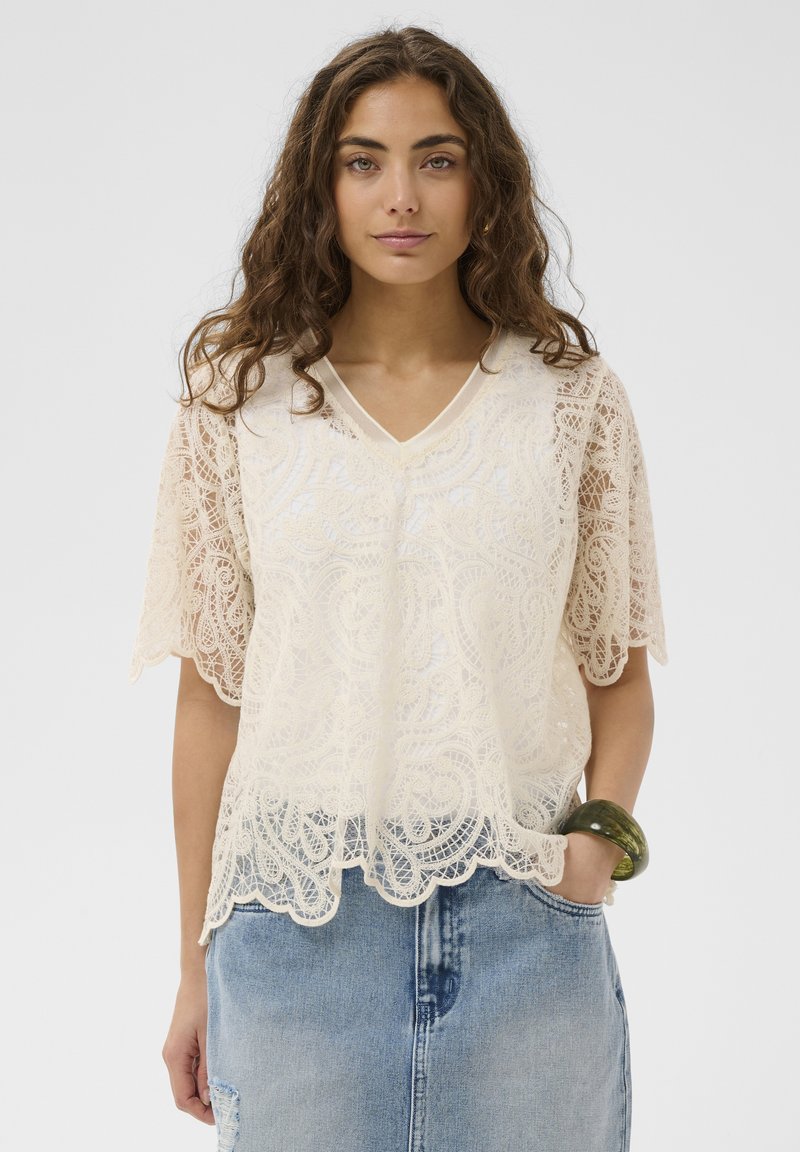 Jeune femme aux cheveux bruns ondulés portant un haut en dentelle crème et une jupe en jean bleu clair, avec un bracelet vert à son poignet.