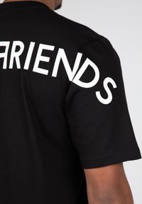 Černé bavlněné tričko s krátkým rukávem, na zadní straně diagonálně potištěné velkým bílým textem "FRIENDS". Hladký povrch, klasický střih.