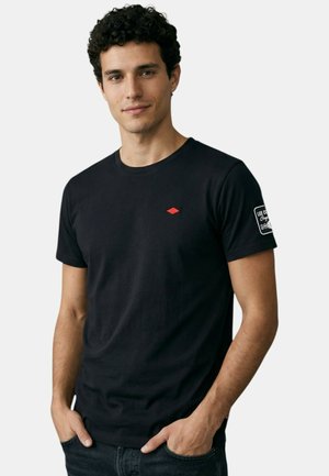 Homme aux cheveux noirs bouclés portant un T-shirt noir avec un petit logo rouge sur la poitrine et un texte blanc sur la manche gauche, les mains dans les poches.