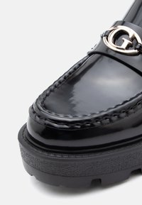 Juodas patentinis odinis loaferis su storesniu guminio pado, detalizuotais siūlais ir išryškintą auksinės spalvos logotipo akcentu ant dirželio.